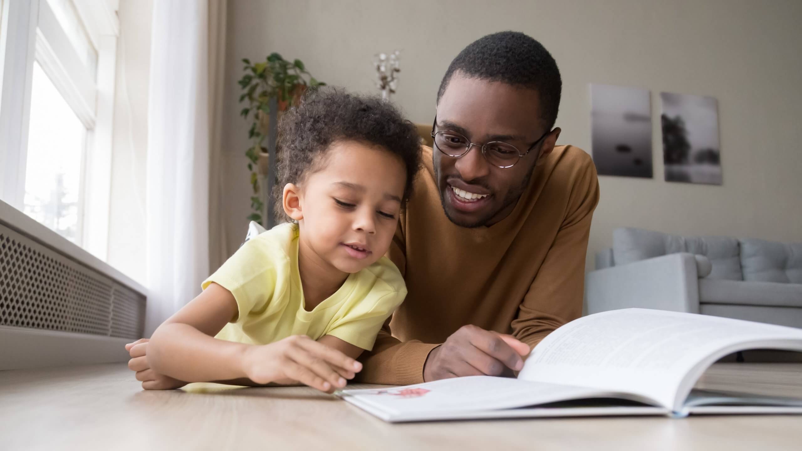 African Father And Son Lying On Warm Floor Reading Book